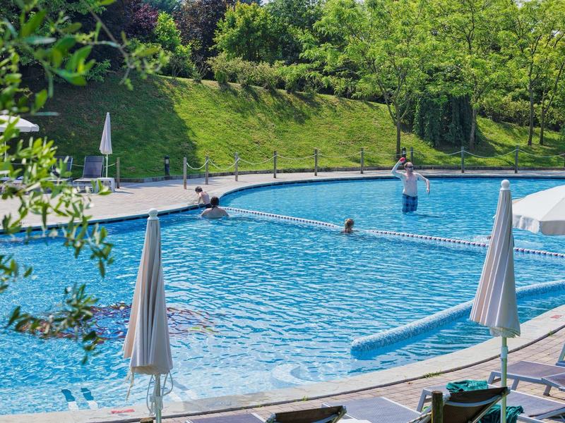 Een buitenzwembad met blauw water, ligstoelen en witte parasols in een groene omgeving.