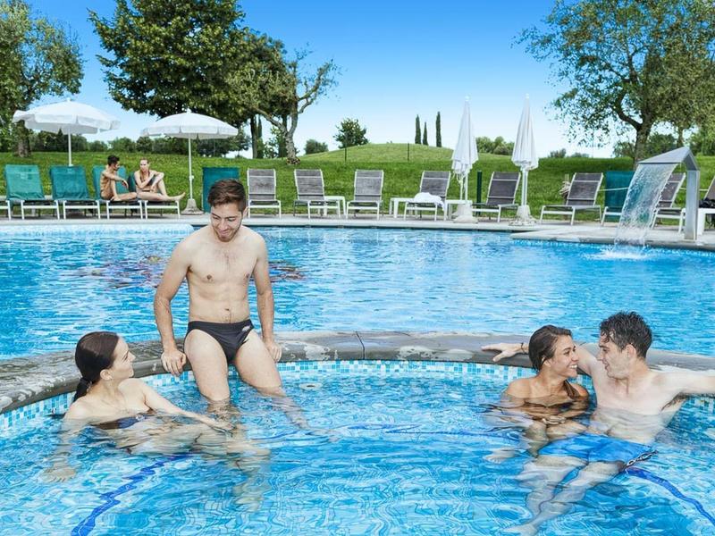 Vier jonge volwassenen genieten van een zomers buitenzwembad met ligstoelen op de achtergrond.