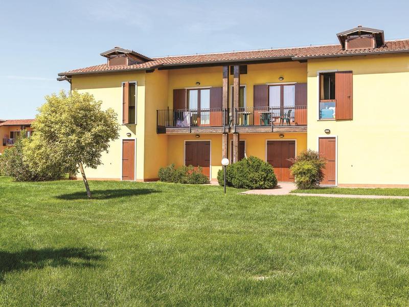 Twee verdiepingen tellend geel vakantiehuis met balkon en groot gazon op de voorgrond.