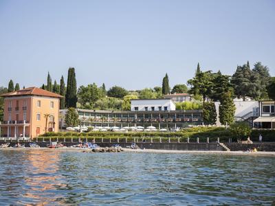 Seeufer mit zwei Gebäuden, grünen Bäumen, Sonnenschirmen und klarem Himmel im Hintergrund.