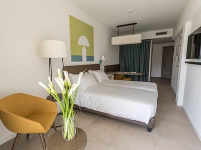 Modern eingerichtetes Hotelzimmer mit Doppelbett, orangem Sessel und großem Blumenstrauß.