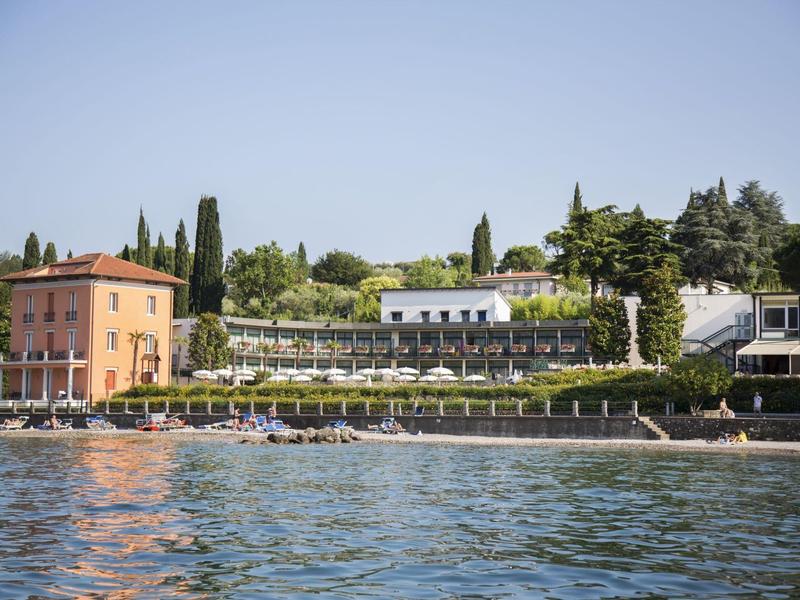 Seeufer mit zwei Gebäuden, grünen Bäumen, Sonnenschirmen und klarem Himmel im Hintergrund.