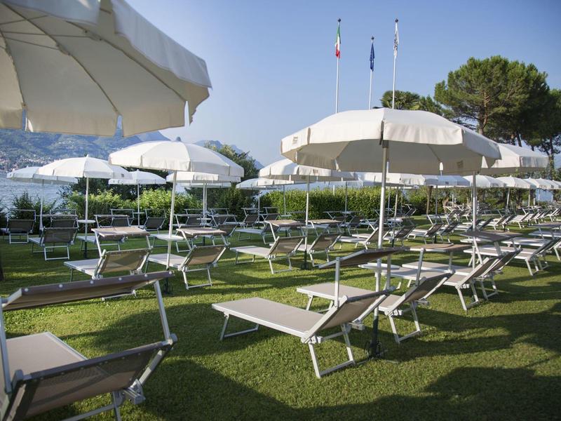 Sonnenliegen und weiße Sonnenschirme stehen geordnet auf einer grünen Wiese bei klarem Himmel.