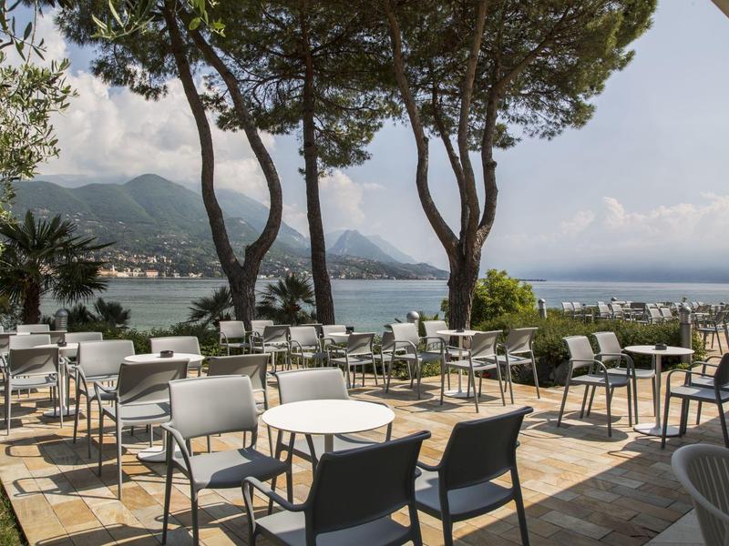 Terrassenbereich mit weißen und schwarzen Stühlen, runden Tischen, Pinienbäumen und Blick auf das Meer.