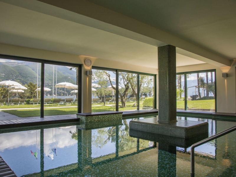 Innenbereich mit großem Schwimmbecken und bodentiefen Fenstern, draußen grüne Wiese und Sonnenschirme.