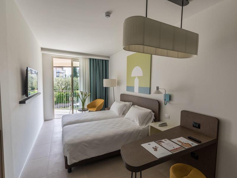 Schlichtes Hotelzimmer mit zwei Betten, Fernseher, kleinem Tisch und Balkon mit Gartenblick.
