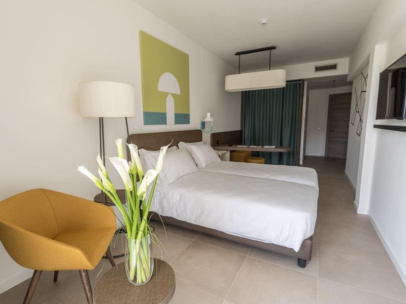 Modern eingerichtetes Hotelzimmer mit Doppelbett, orangem Sessel und großem Blumenstrauß.