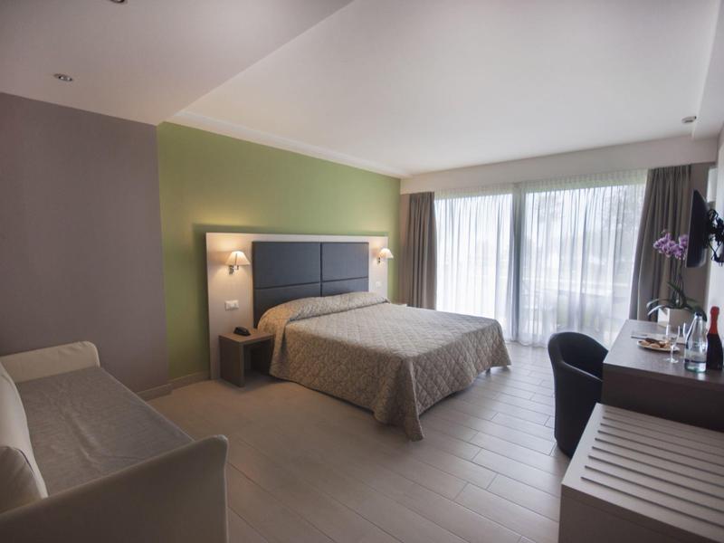 Modernes Hotelzimmer mit Doppelbett, Sofa, Schreibtisch und großem Fenster mit Vorhängen.