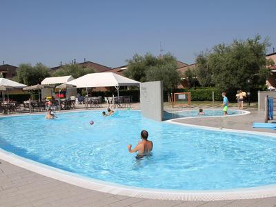 Outdoor pool with people on a sunny day, surrounded by umbrellas and buildings.