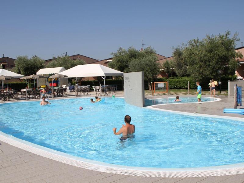 Outdoor pool with people on a sunny day, surrounded by umbrellas and buildings.