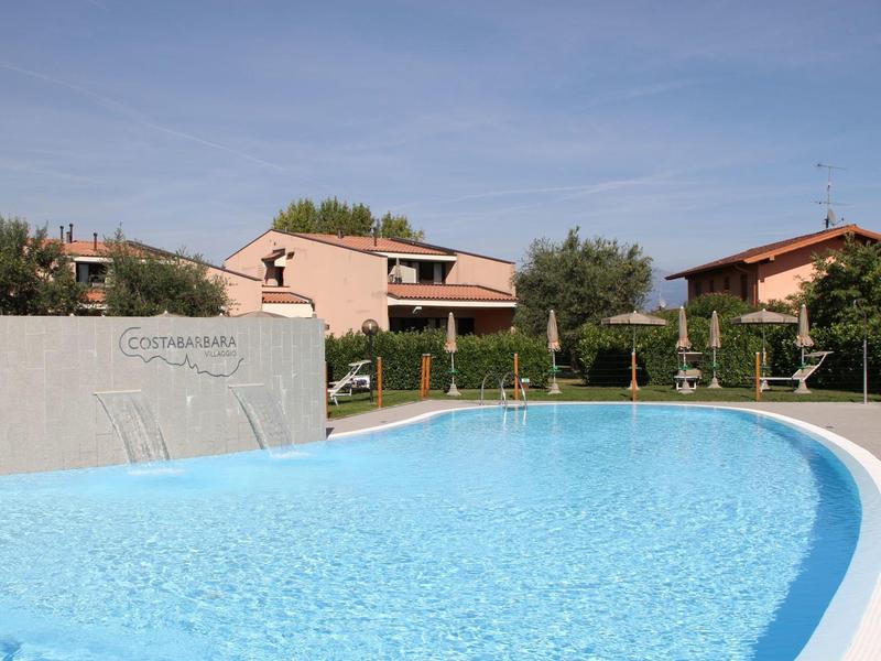 Round outdoor pool in front of several vacation homes with umbrellas under a blue sky.