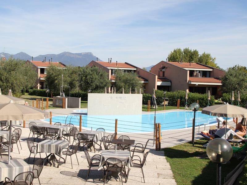 Leisure area with outdoor pool, sun loungers, and tables in front of hotel buildings and mountains.