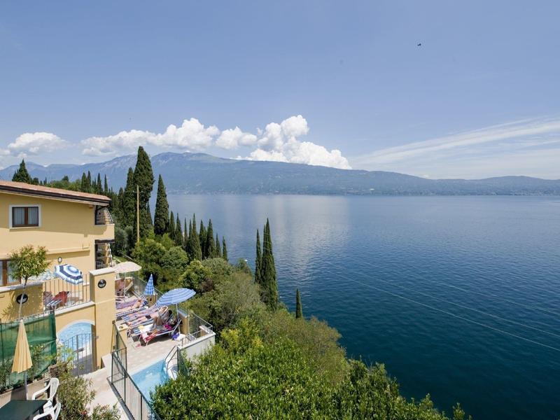 Vista de un lago con montañas, hoteles alrededor y vegetación verde bajo cielo azul.
