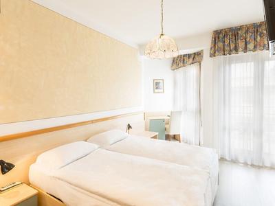 Světý hotelový pokoj se dvěma jednolůžky, stolem, židlí, světlým dekorem a televizí.