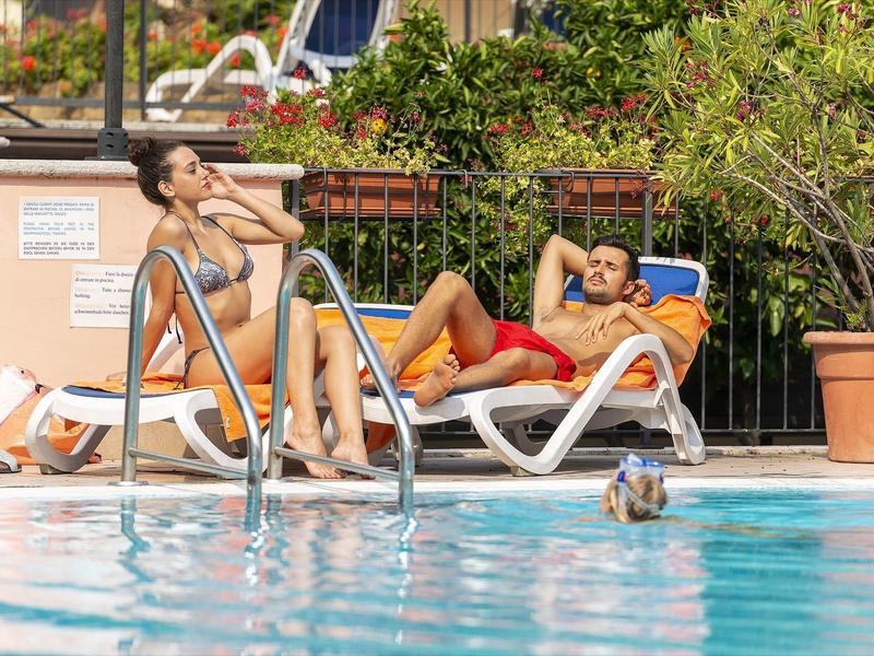 Dva lidé odpočívají na lehátkách vedle bazénu za slunečného počasí.