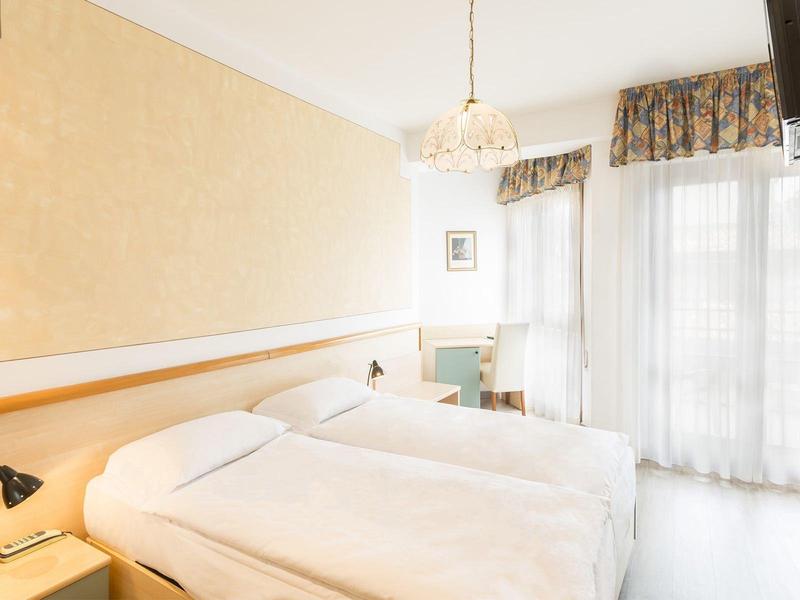 Světý hotelový pokoj se dvěma jednolůžky, stolem, židlí, světlým dekorem a televizí.