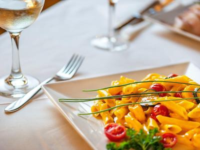 Kieliszek białego wina obok talerza z makaronem i pomidorami na białym obrusie.