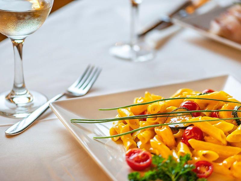 Kieliszek białego wina obok talerza z makaronem i pomidorami na białym obrusie.