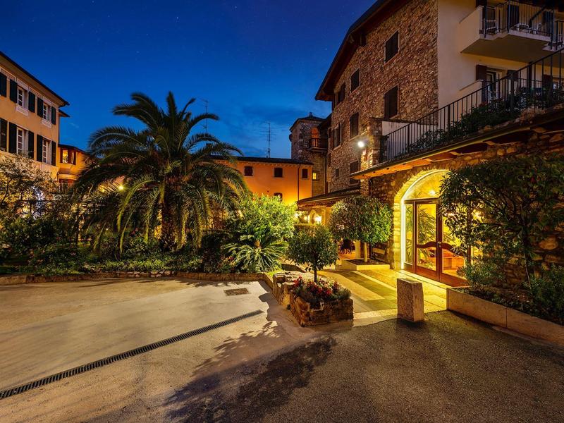 Widok wieczorny jasno oświetlonego hotelu z palmami i brukowanym dziedzińcem.