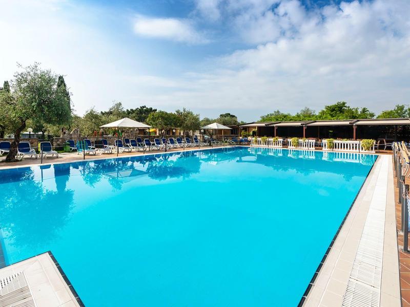 Duży odkryty basen z niebieską wodą i leżakami w słonecznym ogrodzie hotelowym.