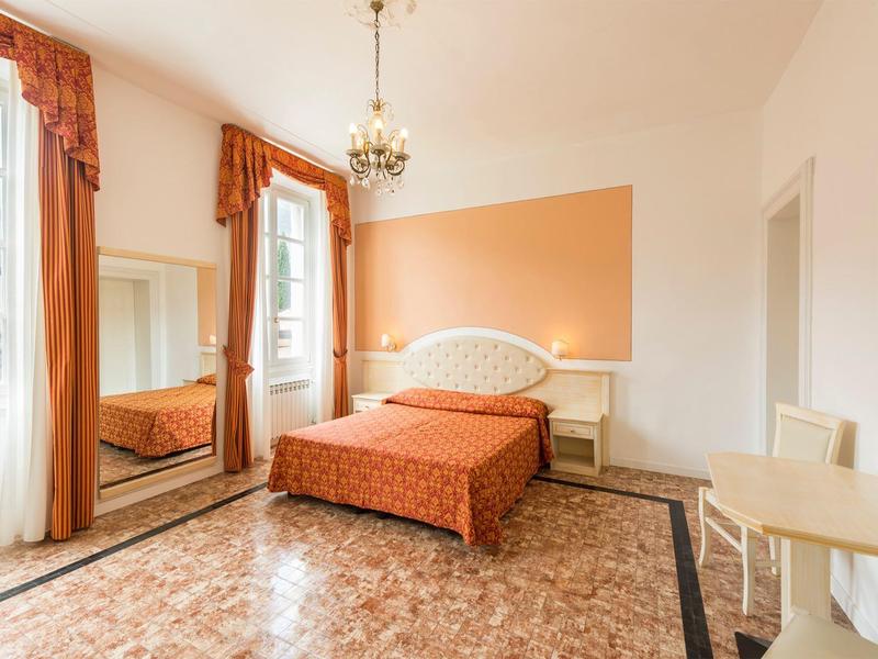 Jasny pokój hotelowy z łóżkiem podwójnym, pomarańczowymi zasłonami i kafelkową podłogą.