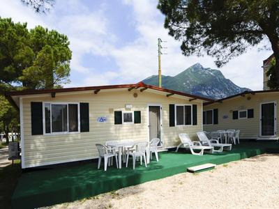 Una casa de vacaciones amarilla con terraza, mesa y sillas, rodeada de árboles y montañas de fondo.