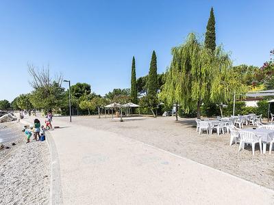 Paseo junto a un playa de guijarros, árboles, áreas de descanso y personas caminando junto al agua.