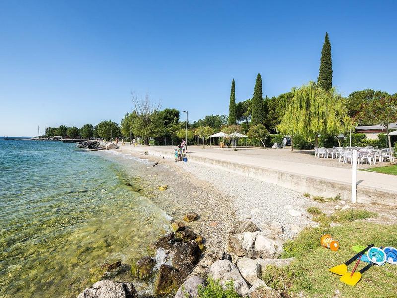 Playa clara con rocas, árboles y pequeños edificios en la orilla bajo un cielo azul.