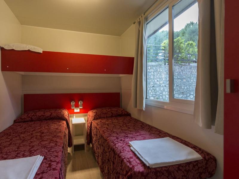 Pequeña habitación de hotel con dos camas individuales, ropa de cama roja y ventana con vista verde.