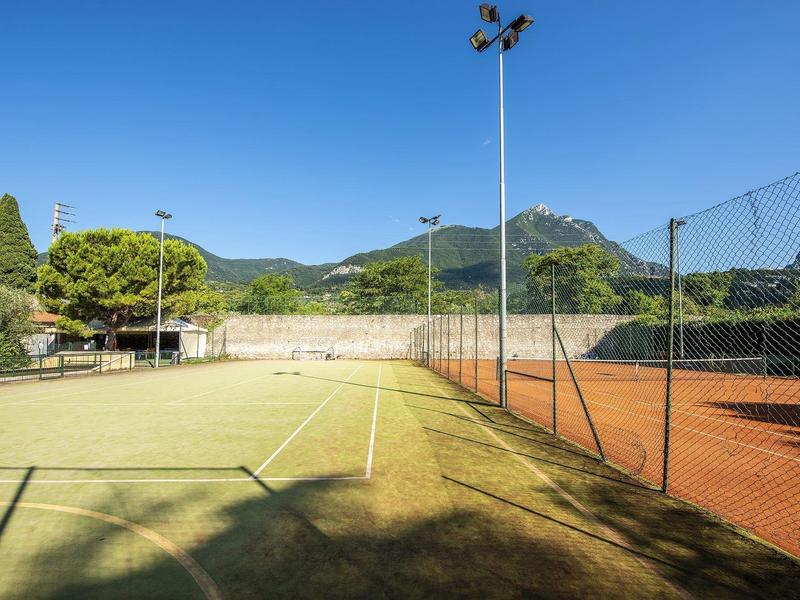 Una cancha de tenis con césped artificial verde junto a una cancha de tierra roja, rodeada de una cerca de alambre y montañas al fondo.