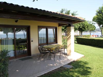 Terrace of a holiday home with table and chairs, surrounded by lawn and overlooking water.