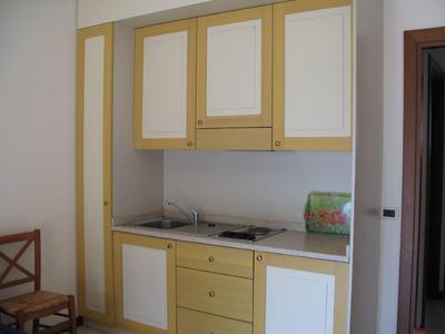 Compact kitchenette with sink, stove, upper cabinets, and chair in neutral colors.