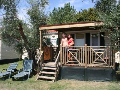 Zwei Personen auf Holzveranda eines kleinen Hauses mit grüner Wiese und Bäumen im Hintergrund.