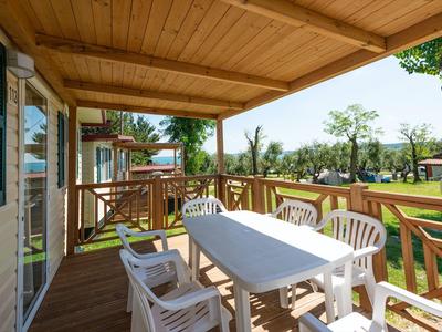 Überdachte Terrasse mit weißem Tisch und Stühlen, Holzboden, grüner Garten und Bäume im Hintergrund.