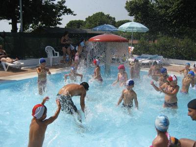 Viele Kinder und ein Erwachsener spielen im Wasser eines Schwimmbeckens bei sonnigem Wetter.