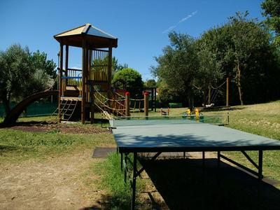 Spielplatz mit Kletterturm, Rutsche und Tischtennisplatte unter blauem Himmel und Bäumen.
