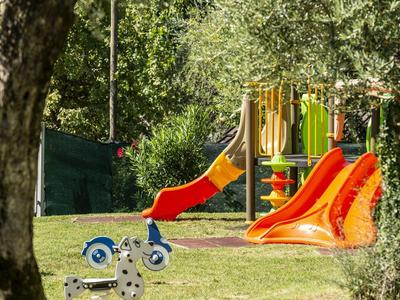 Spielplatz mit bunten Rutschen und Schaukelpferden auf grüner Wiese, umgeben von Bäumen.