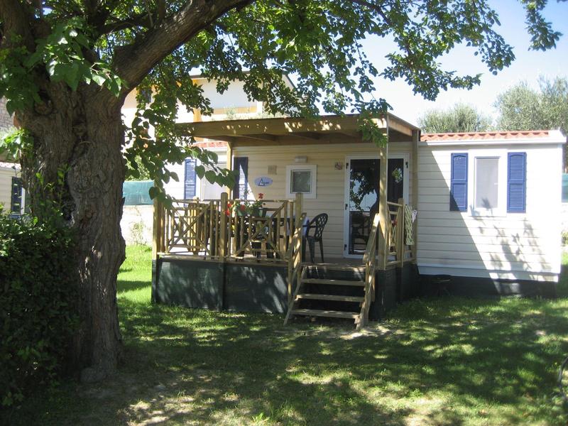 Mobilheim mit Veranda unter großen Bäumen, Holzgeländer und Stufen, im sonnigen Garten.