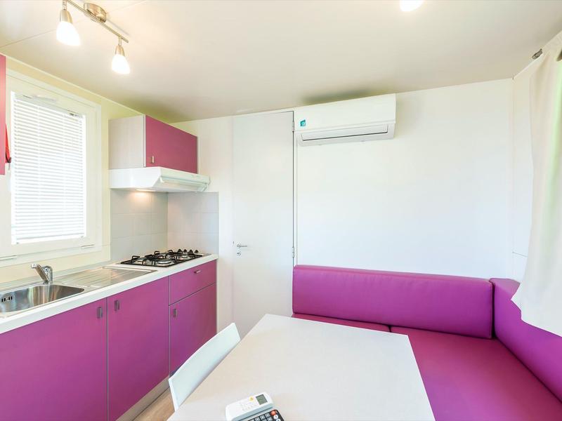 Kleine Küche mit lila Schränken, weißer Tischplatte, Eckbank und Klimagerät an der Wand.