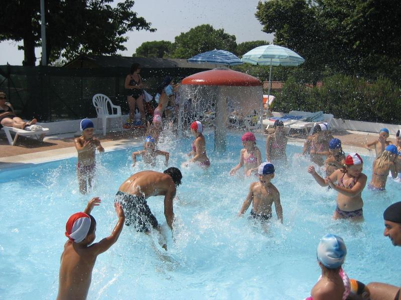 Viele Kinder und ein Erwachsener spielen im Wasser eines Schwimmbeckens bei sonnigem Wetter.