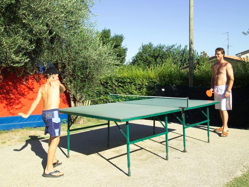 Zwei Männer spielen im Freien Tischtennis auf einem grünen Tisch, umgeben von Büschen und Bäumen.