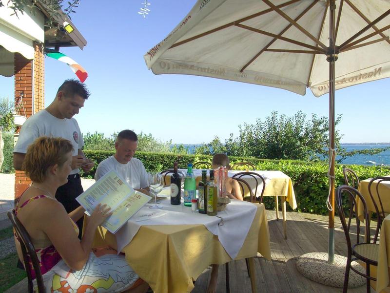 Menschen sitzen an einem Gartentisch mit Sonnenschirm, umgeben von Pflanzen und Meerblick.