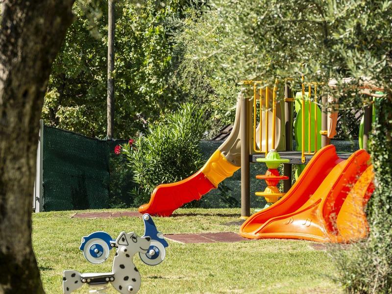 Spielplatz mit bunten Rutschen und Schaukelpferden auf grüner Wiese, umgeben von Bäumen.