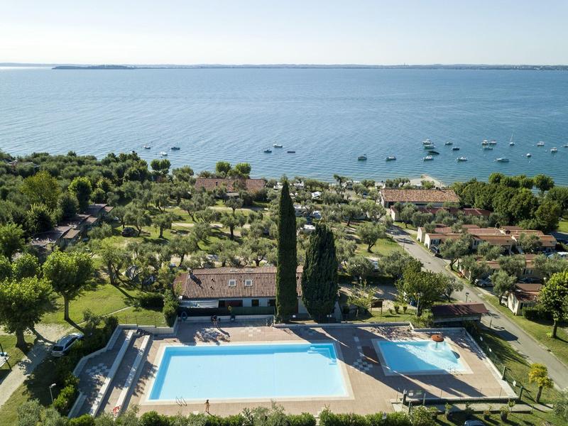 Landschaft mit zwei großen Pools, grünem Garten und vielen Booten auf ruhigem blauen See im Hintergrund.