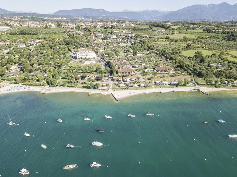 Blick auf eine Küstenstadt mit grünem Land, Strand, Booten im blauen Wasser und Bergen im Hintergrund.