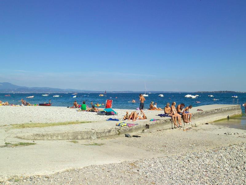 Strand mit Sand, Kies, Menschen, bunten Sonnenschirmen und klarem blauem Himmel.