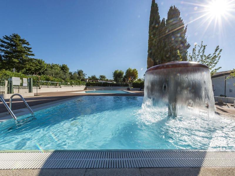 Schwimmbad mit klarem Wasser, Wasserfall und Sonnenschein vor blauem Himmel und Bäumen.