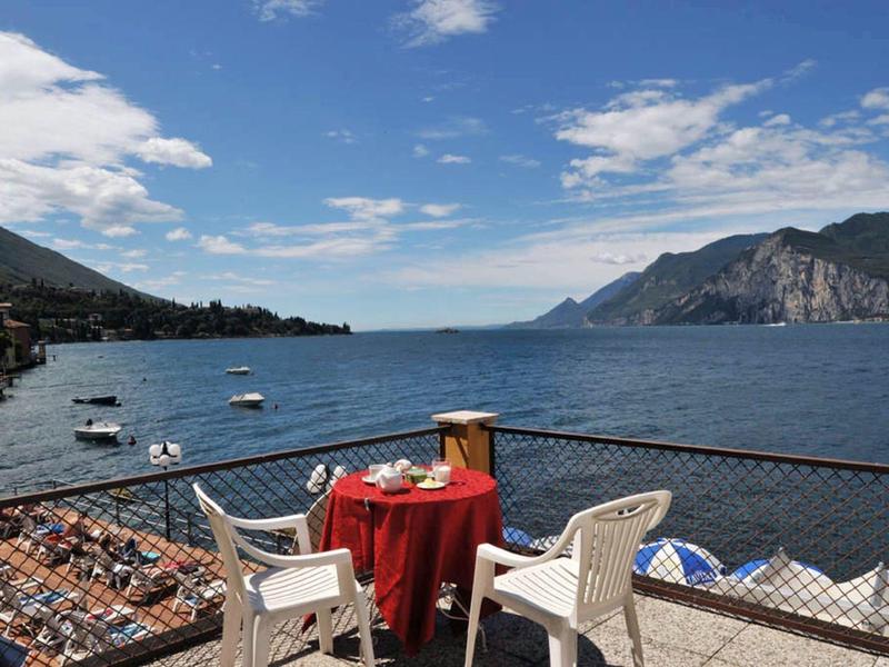 Vista da una terrazza con tavolo e sedie su un lago tranquillo e montagne sullo sfondo.