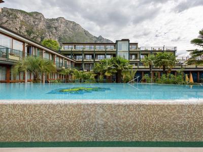 Moderner Hotelpool mit klar blauem Wasser, umgeben von Palmen und Gebäuden unter bewölktem Himmel.