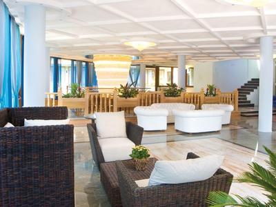 Helles Hotel-Lounge mit weißen Sofas, Rattanstühlen und grünen Pflanzen unter weißer Decke.